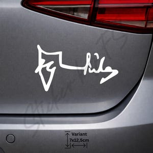 Op de afbeelding: Witte vinyl auto sticker met gestileerd schrift op een grijze autobumper. De tekst luidt "Variant" met afmetingen van 7x12,5 cm.