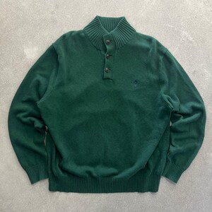 May include: A dark green long-sleeve sweater featuring a ribbed mock neck collar with a three-button placket. The knit garment includes ribbed cuffs and a ribbed hem, with a small dark embroidered logo on the left chest area of the front panel.