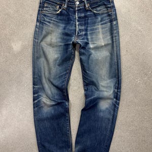 Puede incluir: Unos vaqueros de mezclilla azul oscuro con un lavado desgastado, que cuentan con cierre de botones, cinco bolsillos y remaches de color cobre. La tela muestra patrones de desgaste en las caderas y los muslos, con dobladillos vueltos.
