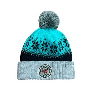 May include: A gray, turquoise, and black knit beanie with a gray pom-pom. The turquoise section features a black snowflake pattern. The gray brim has a circular emblem with a Native American design in red, black, and white.