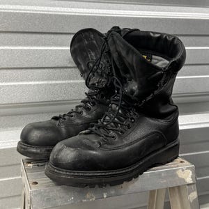 Könnte beinhalten: Ein Paar schwarze taktische Stiefel aus Leder mit hohem Schaft, schwarzen Schnürsenkeln, Metallösen und einer dicken, robusten Gummisohle. Die Stiefel haben einen unteren Bereich aus strukturiertem Leder und einen glatten, flexiblen Schaftabschluss.