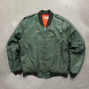 Puede incluir: Una chaqueta tipo bomber de color verde oliva con cuello, puños y dobladillo acanalados en marrón oscuro. La prenda cuenta con cierre de cremallera frontal, dos bolsillos laterales con botones a presión, charreteras, un bolsillo utilitario en la manga izquierda y un forro de color naranja brillante.