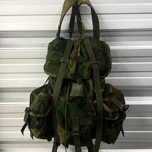 Könnte beinhalten: Ein Rucksack mit Waldtarnmuster, der ein großes Hauptfach mit oberer Klappe, zwei Seitentaschen und eine mittlere Vordertasche aufweist. Der Rucksack verfügt über verstellbare olivgrüne Nylongurte, Kunststoffschließen und ein Kordelzugsystem.