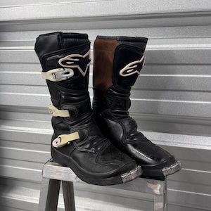 Peut inclure: Une paire de bottes de motocross Alpinestars noires avec des accents en daim marron sur le haut, des boucles en plastique blanc et des embouts métalliques argentés. Les bottes ont une coupe haute avec des panneaux de protection et le logo Alpinestars.