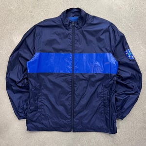 May include: A navy blue windbreaker jacket featuring a horizontal bright blue stripe across the chest, a full-length front zipper, a stand-up collar, and a small circular patch on the left sleeve with white star graphics and blue text.