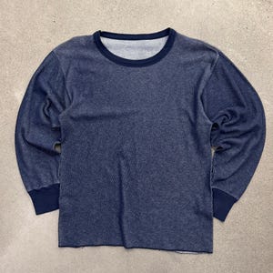 May include: A long-sleeve thermal shirt in a deep navy blue colour. The garment features a textured waffle-knit fabric throughout the body and sleeves, with solid navy blue ribbed trim at the crew neckline and at the cuffs of the long sleeves.