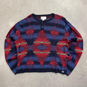 May include: A long-sleeve henley sweater featuring a dark blue, red, and black geometric pattern. The knit garment has a round neckline with a two-button placket, ribbed cuffs, and a hem. A small rectangular brand label is visible at the bottom left.