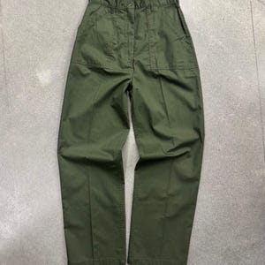 Puede incluir: Pantalones de talle alto en color verde oliva con cierre de botón y cremallera, dos grandes bolsillos rectangulares de parche en la parte delantera, trabillas para cinturón y corte de pierna recta con un pliegue vertical visible en cada pierna.