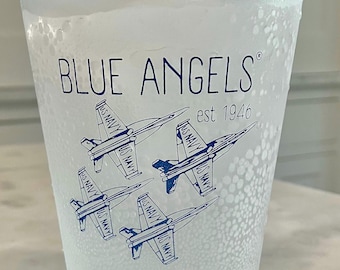 May include: A clear plastic cup with condensation on the outside. The cup has a blue and white design with the words "BLUE ANGELS" and "est 1946". There are four blue and white fighter jets on the cup.