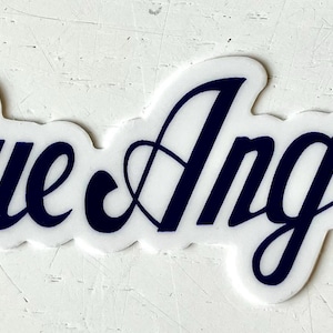 May include: A white sticker with the words "Blue Angels" in a navy blue script font. The sticker has a scalloped edge and a registered trademark symbol.