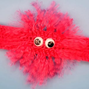 Può includere: Un cerchietto per capelli in crochet rosa acceso con un pompon rosa soffice e due grandi occhietti tondi neri e bianchi.