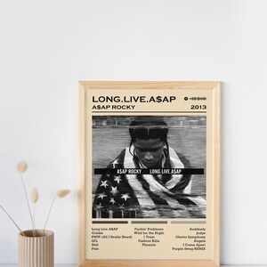 May include: A black and white album cover with a portrait of A$AP Rocky in the center. The album title "LONG.LIVE.A$AP" and the artist's name "A$AP ROCKY" are printed in black text. The album cover is framed in a light wood frame. The album cover also includes a list of songs: "Long Live ASAP", "Goldie", "PMW (All I Really Need)", "LVL", "Hell", "Pain", "Fuckin' Problema", "Wild for the Night", "1 Train", "Fashion Killa", "Phoenix", "Suddenly", "Jodye", "Ghetto Symphony", "Angels", "1 Come Apart", "Purple Swag REMIX".