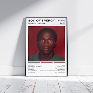 May include: A framed poster featuring a portrait of a person with the text "SON OF SPERGY" and "DANIEL CAESAR" at the top. The poster has a red background and a list of song titles at the bottom. The poster is leaning against a white wall.