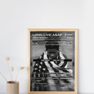 May include: A black and white album cover with a portrait of A$AP Rocky. The album title "LONG.LIVE.A$AP" and the artist's name "A$AP ROCKY" are printed in white text. The album release year "2013" is printed in white text in the top right corner. The album cover features a black and white image of A$AP Rocky wearing a hooded sweatshirt and a bandana. The background of the album cover is a blurred image of an American flag.