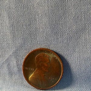 Puede incluir: Una moneda de color cobre con el perfil de una persona en el anverso. La moneda tiene las inscripciones "LIBERTY" e "IN GOD WE TRUST". El año "1988" es visible en la moneda. Fondo azul.