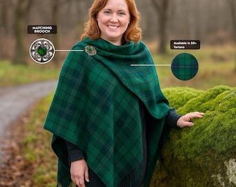 May include: A green and navy plaid cape with fringe, secured with a matching brooch. The image shows a woman wearing the cape. Text on the image says "Matching Brooch" and "Available in 50+ Tartans."