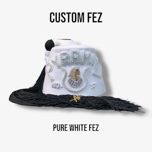 Puede incluir: Un sombrero fez blanco con la palabra "PYRAMID" en pedrería plateada. El sombrero presenta un diseño de temática egipcia, una borla negra y los textos "CUSTOM FEZ" y "PURE WHITE FEZ".