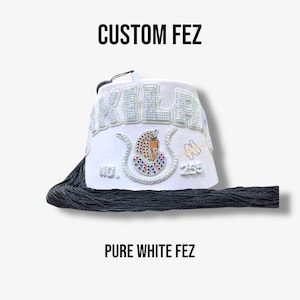 May include: A white fez hat with the text "CUSTOM FEZ" at the top and "PURE WHITE FEZ" at the bottom. The hat features beaded embellishments spelling out a name and a colorful design. A black tassel hangs from the hat.