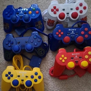 May include: Four different colored Sony Playstation 2 controllers. The controllers are blue, white, red, and yellow. Each controller has the Sony Playstation logo and the words "Playstation" and "Analog" printed on them.