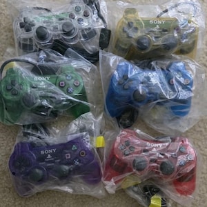May include: Six Sony PlayStation game controllers in individual clear plastic bags. The controllers are translucent and come in various colors including clear, yellow, green, blue, purple, and red, each featuring black buttons and analog sticks.
