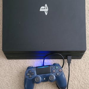 May include: A black PlayStation 4 Pro console with a matching blue controller. The console has the PlayStation logo in white. The controller is connected to the console via a USB cable.