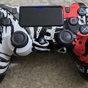 May include: A wireless video game controller featuring a high-contrast graphic design with black, white, and red colors. The left side is white with black abstract patterns, while the right side is red with black splatter and geometric shapes.