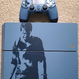 Könnte beinhalten: Eine blaue PlayStation 4 Konsole mit einem blauen Controller. Die Konsole zeigt auf der Vorderseite eine Silhouette eines Mannes mit dem in Gold gedruckten Wort "Uncharted".