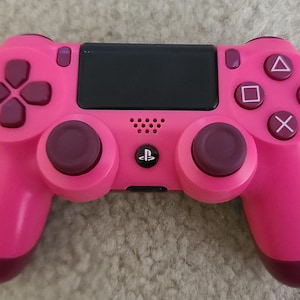 May include: A pink PlayStation 4 DualShock 4 controller with a black faceplate and white buttons. The controller has a pink and purple colour scheme.