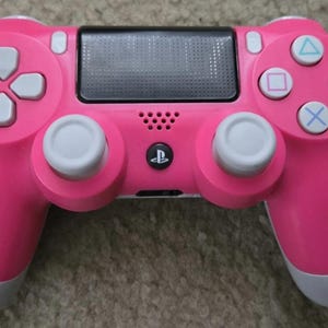 May include: A bright pink video game controller featuring a black central touchpad, two light gray analog sticks, a directional pad, and four action buttons with light blue and pink symbols. A small black PlayStation logo button is centered.