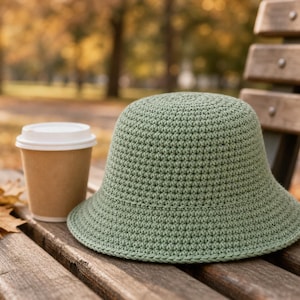 Peut inclure: Un chapeau cloche en crochet vert sauge &agrave; large bord est pos&eacute; sur un banc en bois &agrave; c&ocirc;t&eacute; d'un gobelet de caf&eacute; jetable. Le chapeau est fait d'un fil textur&eacute;. Des feuilles mortes sont &eacute;parpill&eacute;es sur le banc. L'arri&egrave;re-plan est flou.