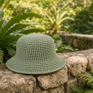 Peut inclure: Un chapeau en crochet vert sauge avec une couronne arrondie et un bord l&eacute;g&egrave;rement &eacute;vas&eacute;. Le chapeau repose sur un mur de pierre, entour&eacute; d'un feuillage vert luxuriant. La texture du chapeau est visible.