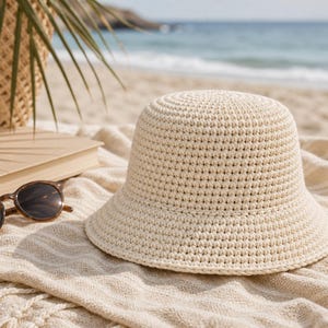 Peut inclure: Un chapeau de seau en crochet cr&egrave;me avec un large bord est pos&eacute; sur une serviette de plage. Des lunettes de soleil &eacute;cailles de tortue et un livre ferm&eacute; sont &agrave; c&ocirc;t&eacute; du chapeau. Un panier tress&eacute; et la mer sont en arri&egrave;re-plan. Le chapeau est un accessoire d'&eacute;t&eacute;.