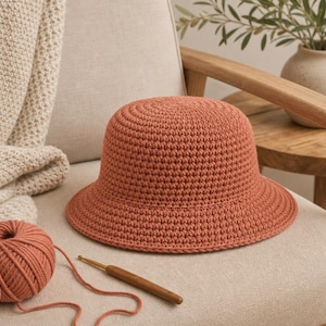 Peut inclure: Un chapeau seau en crochet couleur terre cuite est pos&eacute; sur une chaise, &agrave; c&ocirc;t&eacute; d'une pelote de laine et d'un crochet. Le chapeau a une couronne arrondie et un bord l&eacute;g&egrave;rement &eacute;vas&eacute;. Une couverture en tricot beige est drap&eacute;e sur la chaise.