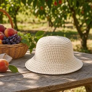 Peut inclure: Un chapeau de soleil en crochet cr&egrave;me avec un bord l&eacute;g&egrave;rement &eacute;vas&eacute; est pos&eacute; sur une table en bois rustique. Un panier en osier rempli de p&ecirc;ches et de raisins est &agrave; proximit&eacute;. Une p&ecirc;che et une feuille sont &eacute;galement sur la table.