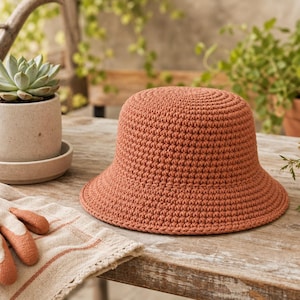 Peut inclure: Un chapeau cloche en crochet couleur terre cuite avec un large bord est pos&eacute; sur une surface en bois. Le chapeau est &agrave; c&ocirc;t&eacute; de gants de jardinage et d'une petite plante succulente dans un pot en c&eacute;ramique. La sc&egrave;ne se d&eacute;roule &agrave; l'ext&eacute;rieur avec de la verdure en arri&egrave;re-plan.