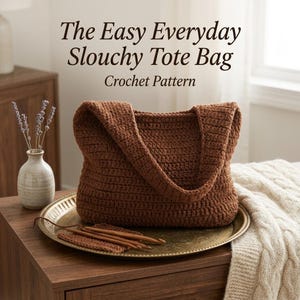 May include: A brown crochet tote bag with a slouchy design, displayed on a gold tray with wooden crochet hooks. The text reads: The Easy Everyday Slouchy Tote Bag Crochet Pattern. The bag features a textured stitch pattern and wide straps.