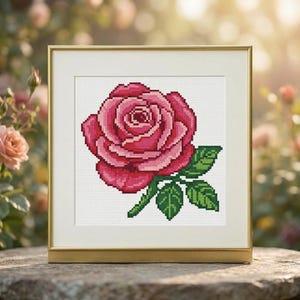 May include: A square gold frame containing a cross-stitch embroidery of a single rose. The rose features layered petals in shades of deep pink and light pink, accompanied by green leaves and a stem, all set against a white grid background.
