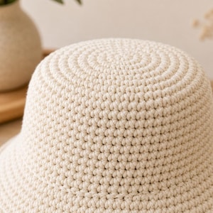 Peut inclure: Gros plan sur un chapeau de p&ecirc;cheur en crochet de couleur cr&egrave;me. Le chapeau pr&eacute;sente une couronne arrondie avec des cercles concentriques et un large bord. La texture des points de crochet est clairement visible, cr&eacute;ant un aspect doux et textur&eacute;. Le chapeau est de couleur neutre.