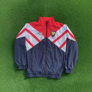 May include: A red, white, and navy blue zip-up windbreaker jacket featuring a high collar, long sleeves with elastic cuffs, and a front zipper closure. The chest displays the text Barry's Tea and a crest with two towers and a shield emblem.