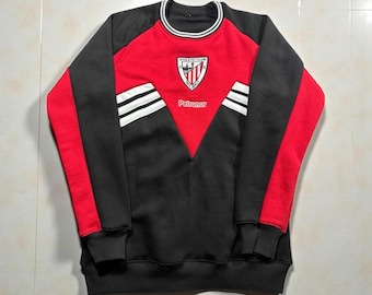 Puede incluir: Una sudadera del Athletic Club roja, negra y blanca. La sudadera presenta una parte superior roja, paneles laterales negros y una sección inferior negra. El logotipo del Athletic Club y "Petronor" son visibles en la parte delantera.