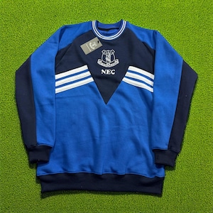 Könnte beinhalten: Ein blaues und marinefarbenes Sweatshirt mit weißen Streifen und einem Wappen-Design. Das Sweatshirt hat einen Rundhalsausschnitt und lange Ärmel. Das Wappen enthält den Text "NEC". Das Sweatshirt ist aus einem weichen Material.