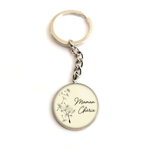 May include: A silver keychain with a round charm featuring a cream background with black dandelion illustrations and the text "Maman Cherie".