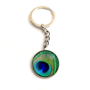 以下が含まれることがあります： 孔雀の羽根のデザインが施された丸いチャームが付いたシルバーのキーチェーン。羽根は緑、青、黄色の色合いで、中央に青い目があります。