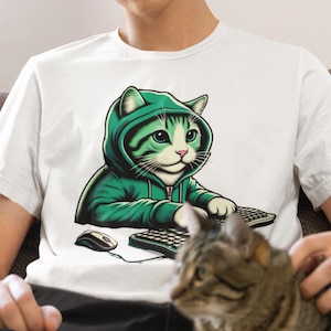 Op de afbeelding: Een wit T-shirt met een afbeelding van een kat die een groene hoodie draagt en op een toetsenbord typt. De kat heeft groene ogen en een witte borst. Het toetsenbord is zwart en heeft witte toetsen. Een muis bevindt zich links van het toetsenbord. Een bruine tabbykat zit op de schoot van de persoon die het shirt draagt.