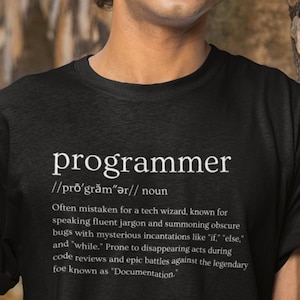May include: Black t-shirt with a white graphic design that defines the word "programmer" with a humorous definition. The definition reads: "Often mistaken for a tech wizard, known for speaking fluent jargon and summoning obscure bugs with mysterious incantations like "if," "else," and "while." Prone to disappearing acts during code reviews and epic battles against the legendary foe known as "Documentation."