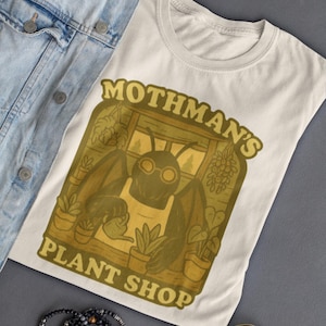 Puede incluir: Camiseta color crema con un diseño vintage de Mothman en una tienda de plantas, con el texto "Mothman's Plant Shop". La camiseta está sobre una superficie gris con una chaqueta vaquera, gafas de sol y pulseras.