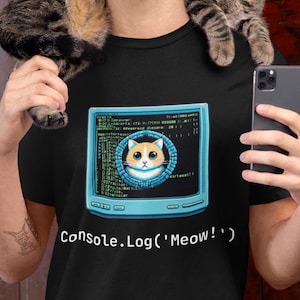 Puede incluir: Una camiseta negra con un gráfico de pantalla de ordenador azul y blanco que presenta la cara de un gato y código. El texto "Console.Log('Meow!')" está impreso debajo del gráfico.