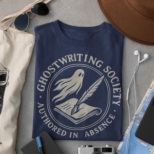 May include: A navy blue t-shirt with a white circle graphic featuring a ghost writing in a book with a quill pen. The text around the circle reads "Ghostwriting Society. Authored in Absence."