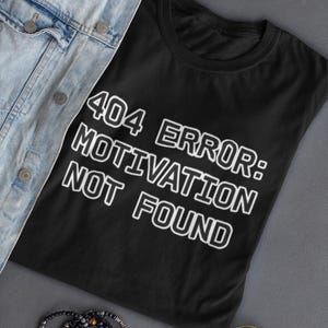 Könnte beinhalten: Schwarzes T-Shirt mit dem weißen Text "404 ERROR: MOTIVATION NOT FOUND". Das Shirt liegt neben einer hellblauen Jeansjacke, einem Perlenarmband und einer goldgerahmten Sonnenbrille.