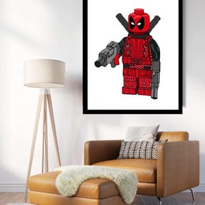 May include: A framed print of a red and black Lego minifigure of Deadpool. The minifigure is holding a gun and has a sword on its back. The background is white.